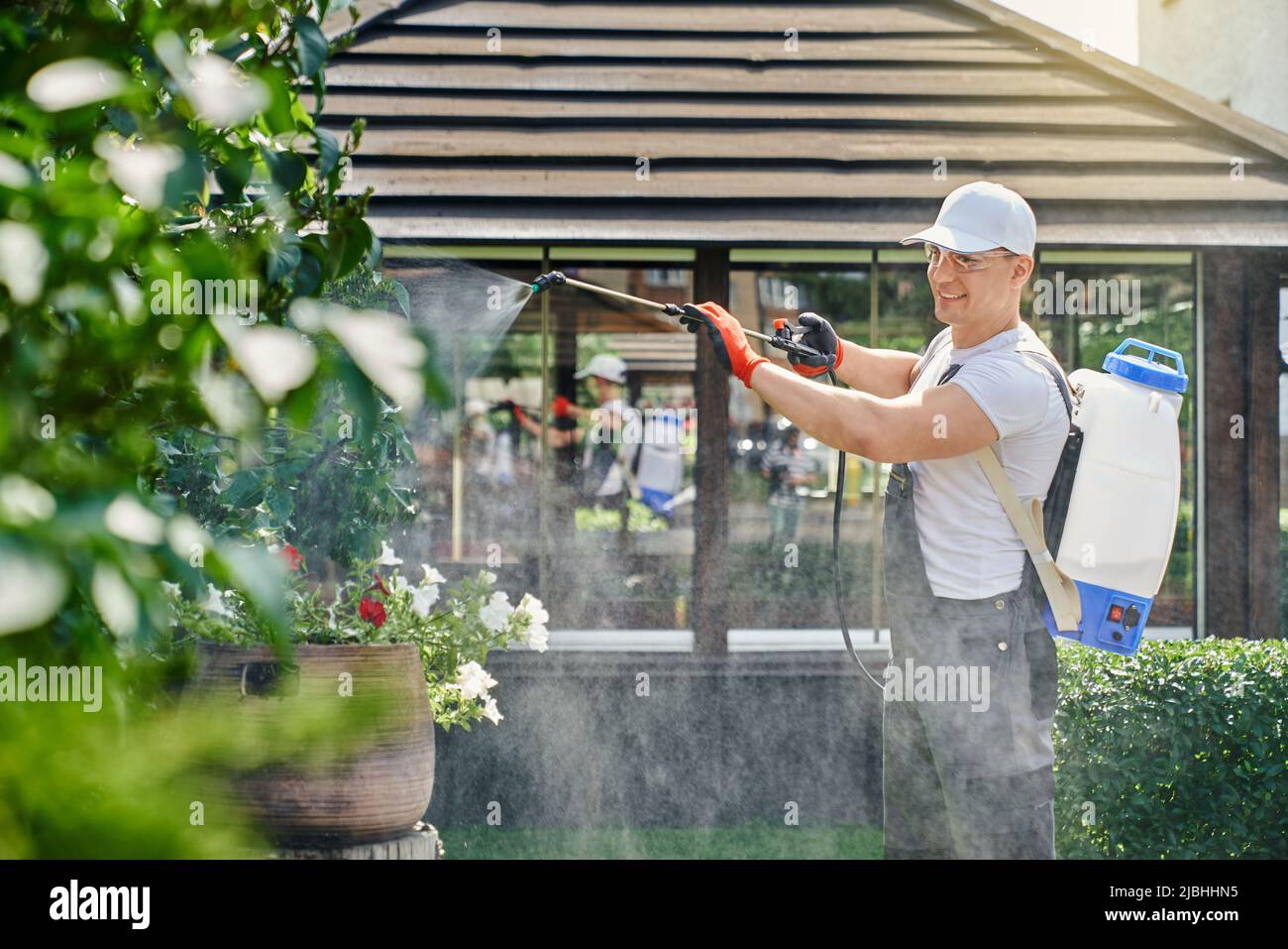 gardner competente in indumenti protettivi, cappello, occhiali e guanti con pulverizzatore per spruzzare piante verdi con prodotti chimici. Uomo caucasico che si prende cura di bel giardino durante la stagione estiva. Foto Stock