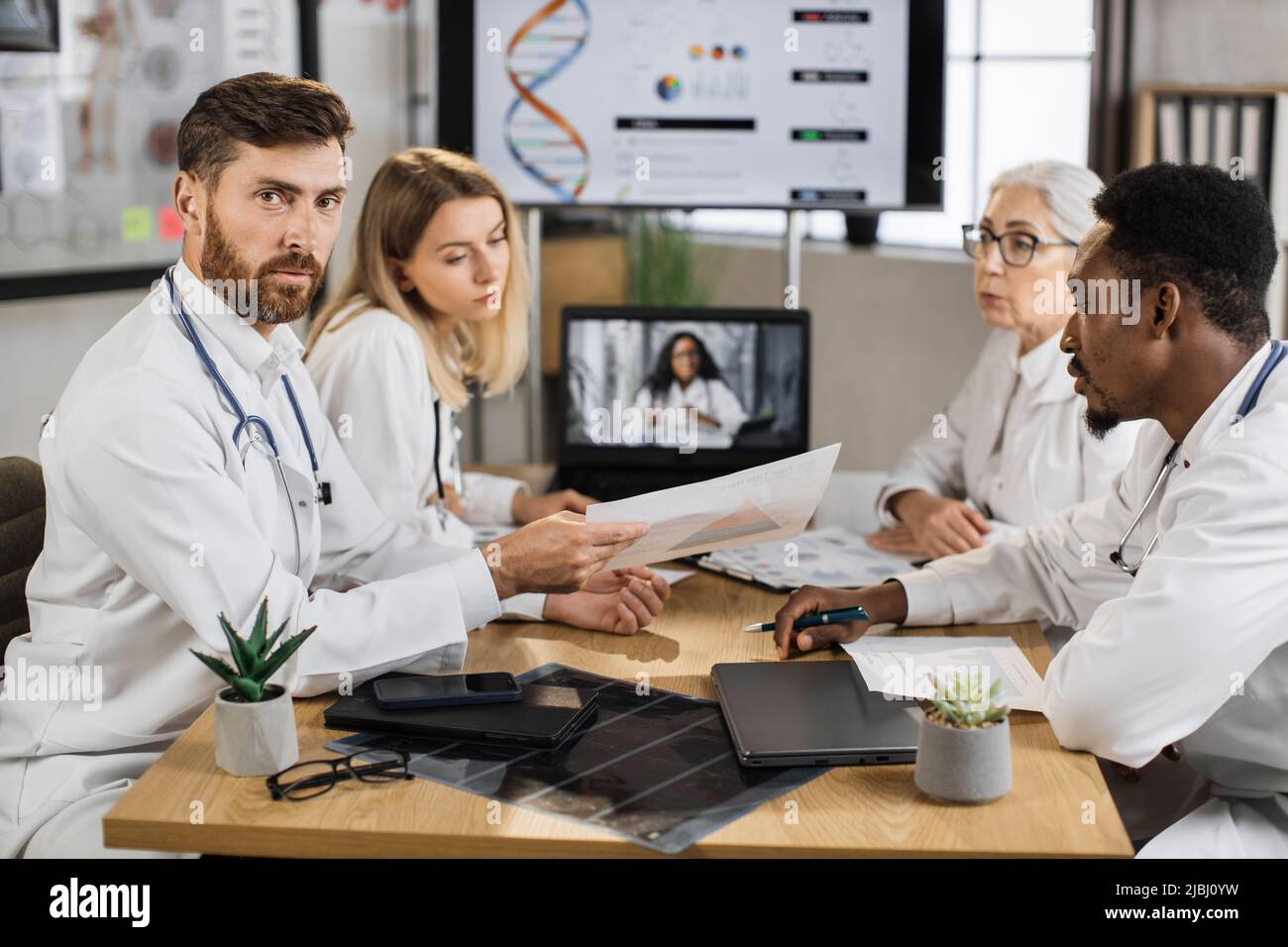 Medico caucasico maschio guardando la macchina fotografica mentre si siede alla scrivania con la presentazione elenco in mani. Colleghi multirazziali seduti vicino e ascoltare la donna afroamericana durante la videochiamata sul laptop. Foto Stock