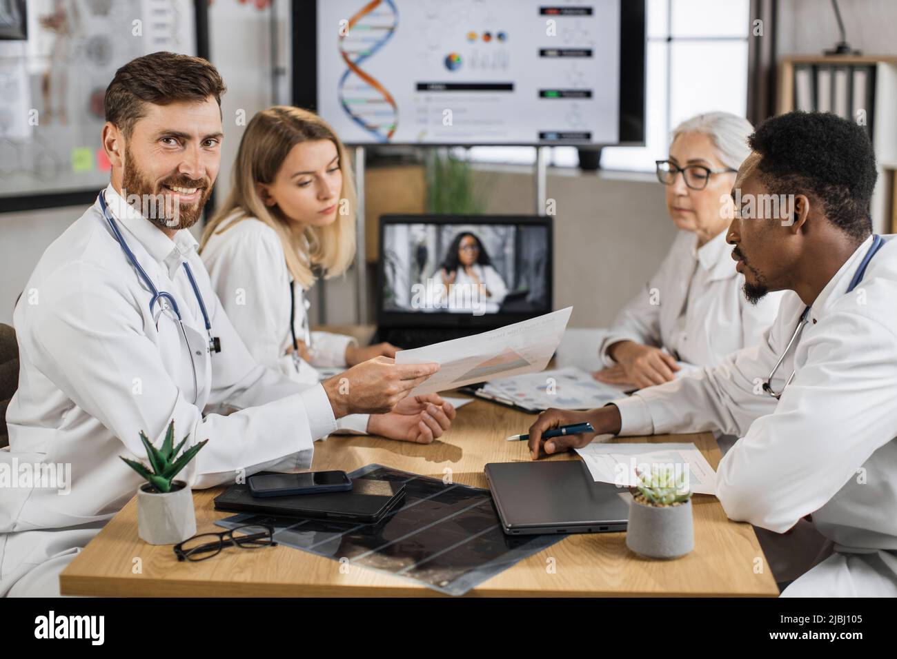 Medico caucasico maschio guardando la macchina fotografica mentre si siede alla scrivania con la presentazione elenco in mani. Colleghi multirazziali seduti vicino e ascoltare la donna afroamericana durante la videochiamata sul laptop. Foto Stock