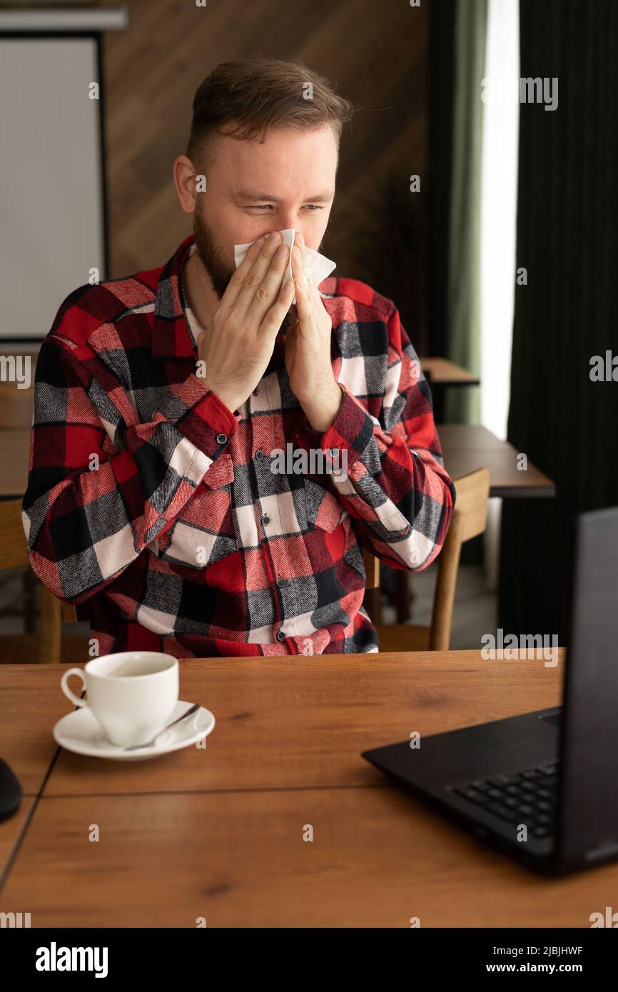 L'uomo caucasico malato che guarda lo schermo del laptop e starnutisce il naso in carta, seduto in ufficio, spazio vuoto. Malattia sinusitica, raffreddore e. Foto Stock