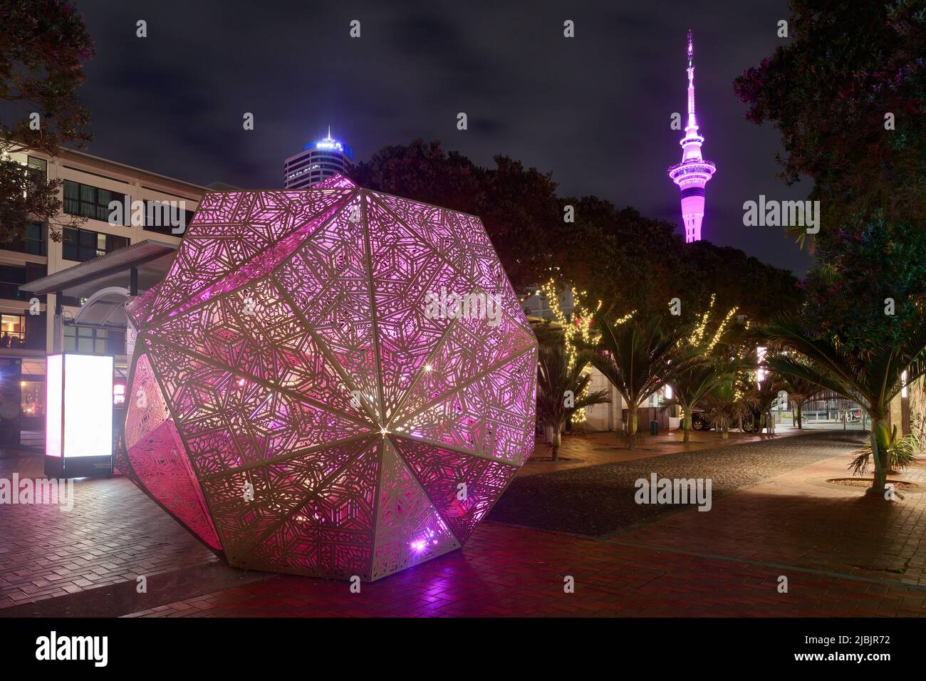 Auckland, Nuova Zelanda. Una scultura geometrica illuminata chiamata 'pensiero dorso' nell'area di Viaduct Harbour, con la Sky Tower sullo sfondo Foto Stock