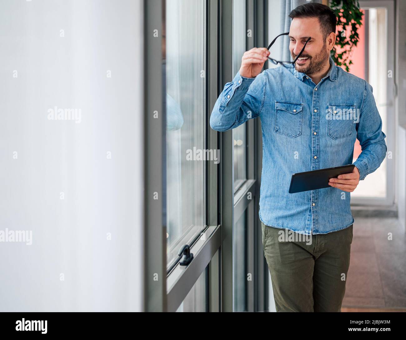Uomo d'affari sorridente con tablet digitale che indossa occhiali. Maschio professionista sta guardando attraverso la finestra. Indossa astuti casuals in ufficio. Foto Stock