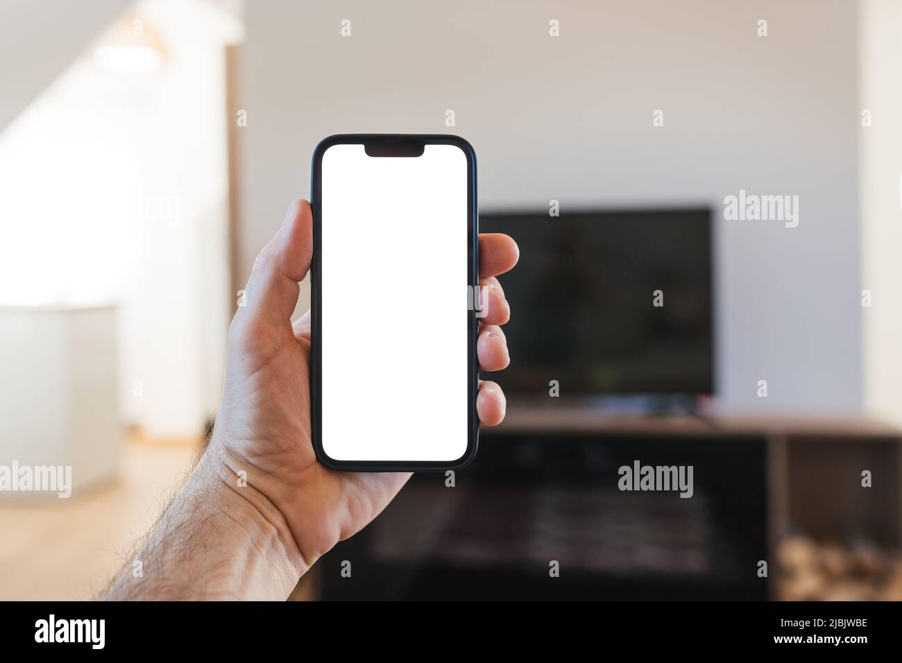 Uomo che tiene smartphone con bianco bianco bianco mockup schermo di fronte al televisore a schermo piatto, messa a fuoco selettiva Foto Stock