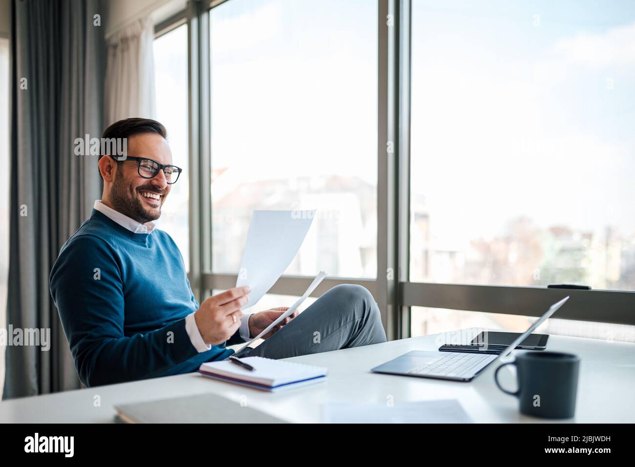 Sorridente professionista aziendale che analizza i documenti. Il dirigente maschile indossa casuals intelligenti. È seduto alla scrivania in ufficio aziendale. Foto Stock