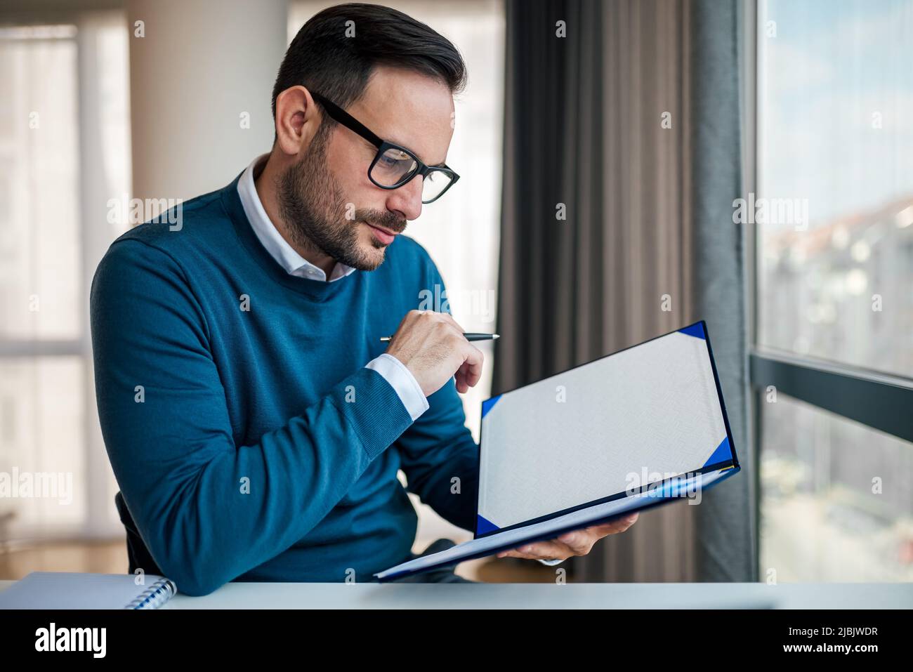 Un giovane uomo d'affari concentrato che analizza i documenti. Il professionista serio sta indossando i casuals astuti. Lavora alla scrivania in ufficio. Foto Stock
