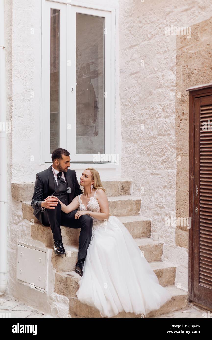 Ritratto di giovane sposa bionda in abito bianco e brunet sposo in vestito guardando l'un l'altro e seduto su scale di pietra vicino al vecchio edificio della città Foto Stock