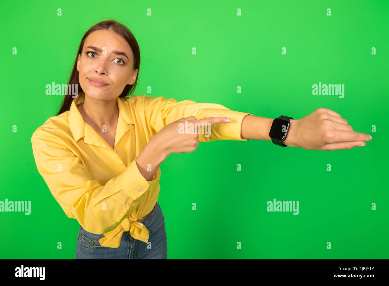 Donna scontenta puntando dito sullo smartwatch in piedi su sfondo verde Foto Stock