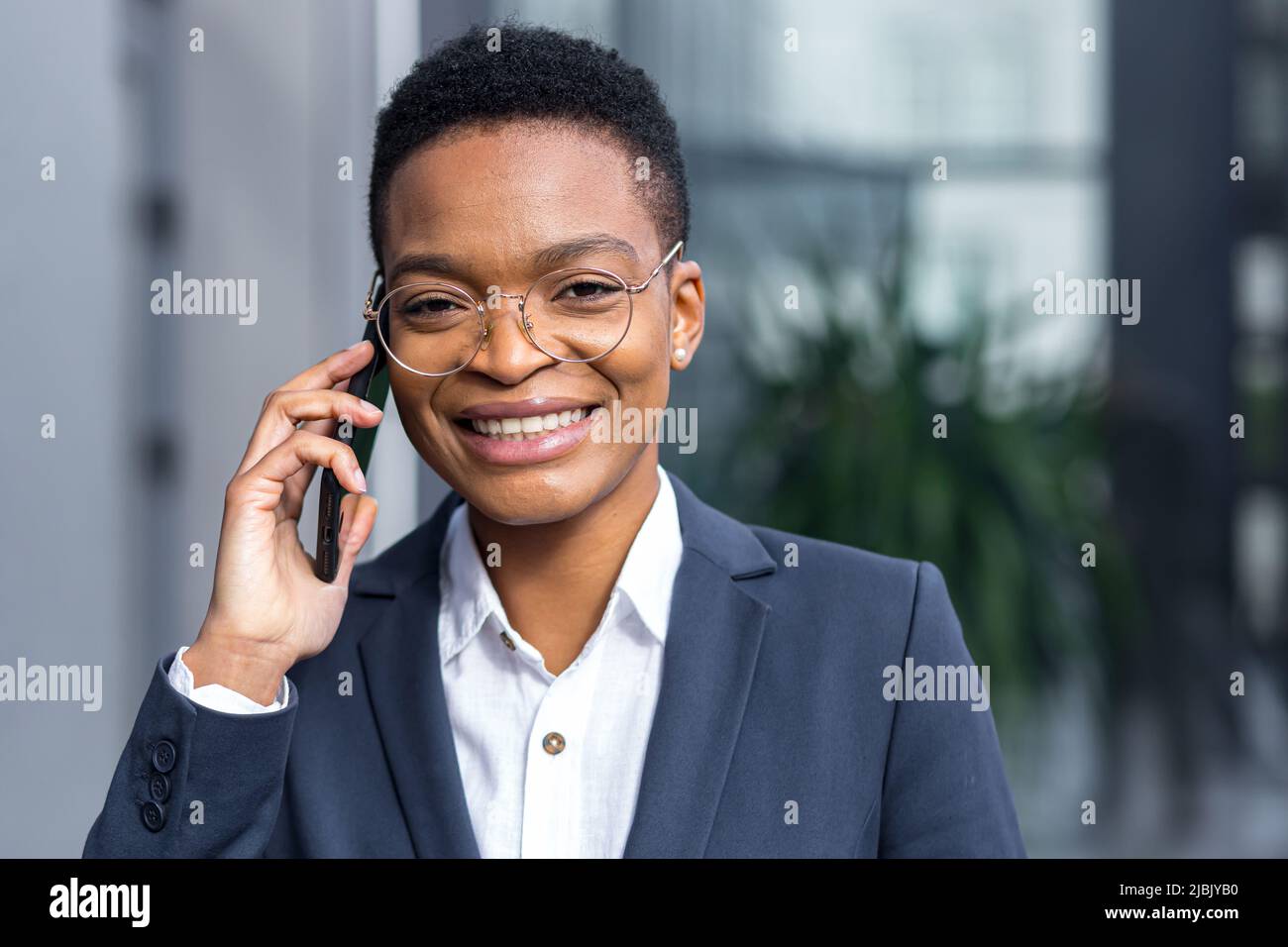 Foto di primo piano ritratto di giovane bella donna da lavoro, afroamericana che parla al telefono, sorridente e rallegrante guardando felice camer Foto Stock