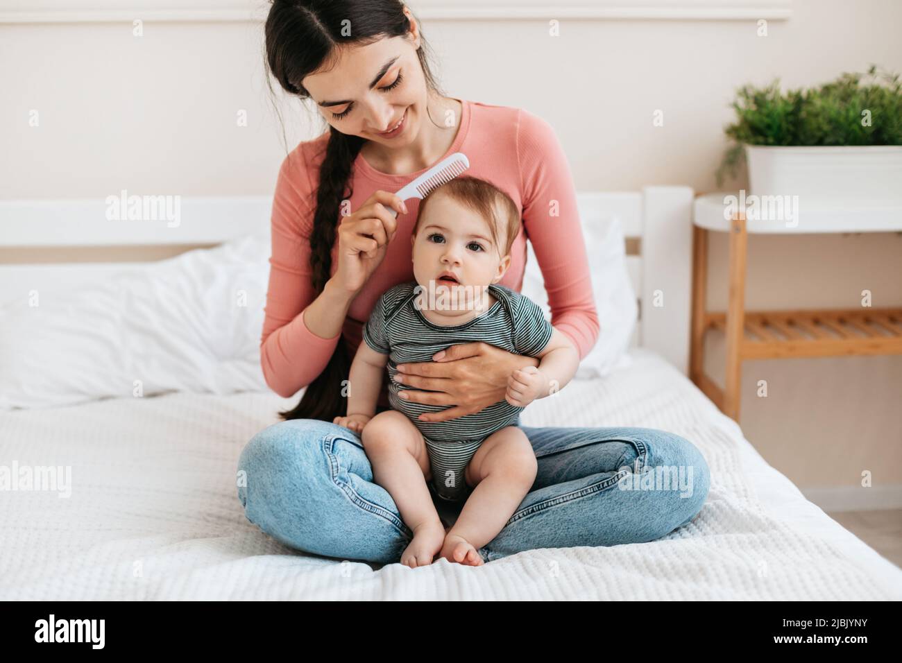 La madre che si prende cura di spazzolare i capelli della figlia, il bambino seduto sui giri della mamma e guardando la macchina fotografica, la donna che ama l'infanzia Foto Stock