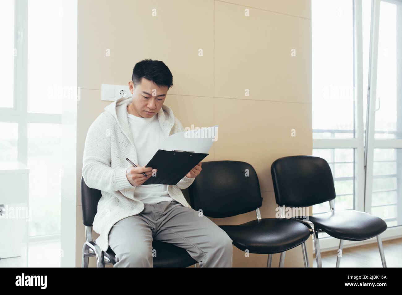 il giovane maschio asiatico in sala d'attesa per l'intervista o riunione tenendo carta mentre si siede alla sedia compila un questionario curriculum in ufficio Foto Stock