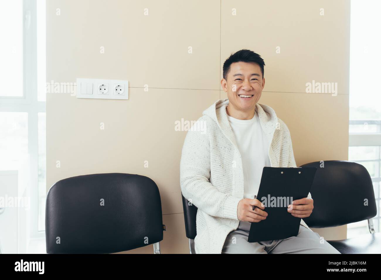 il giovane maschio asiatico in sala d'attesa per l'intervista o riunione tenendo carta mentre si siede alla sedia compila un questionario curriculum in ufficio Foto Stock