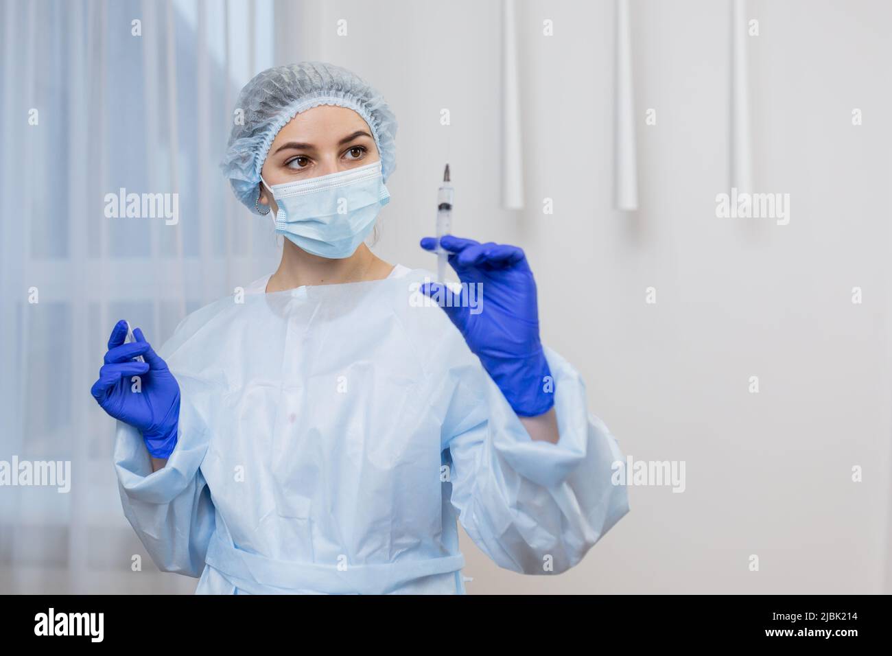 Ritratto di giovane medico infermiere femmina in cappotto bianco in maschera fessura cappuccio chirurgico e guanti medici blu che reggono la siringa, guardando la fotocamera Foto Stock