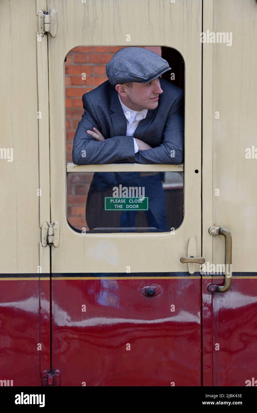 Un uomo in abiti vintage che si stende da una finestra carrozza su un treno a vapore storico a Bewdley Station, Severn Valley Railway, Regno Unito Foto Stock