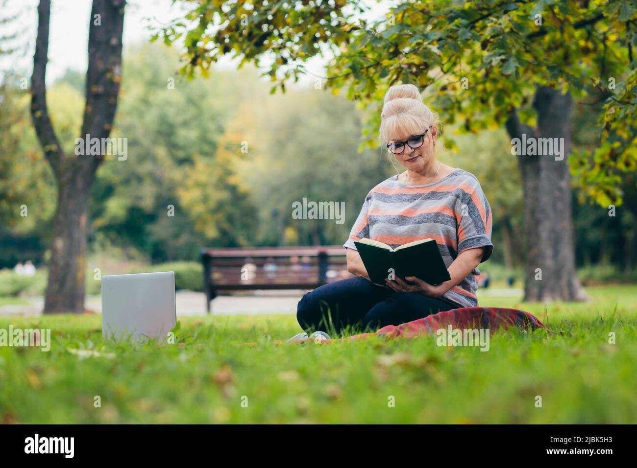 Donna bionda senior bella seduta sul prato nel parco legge un libro, studi, sorrisi, riposa Foto Stock