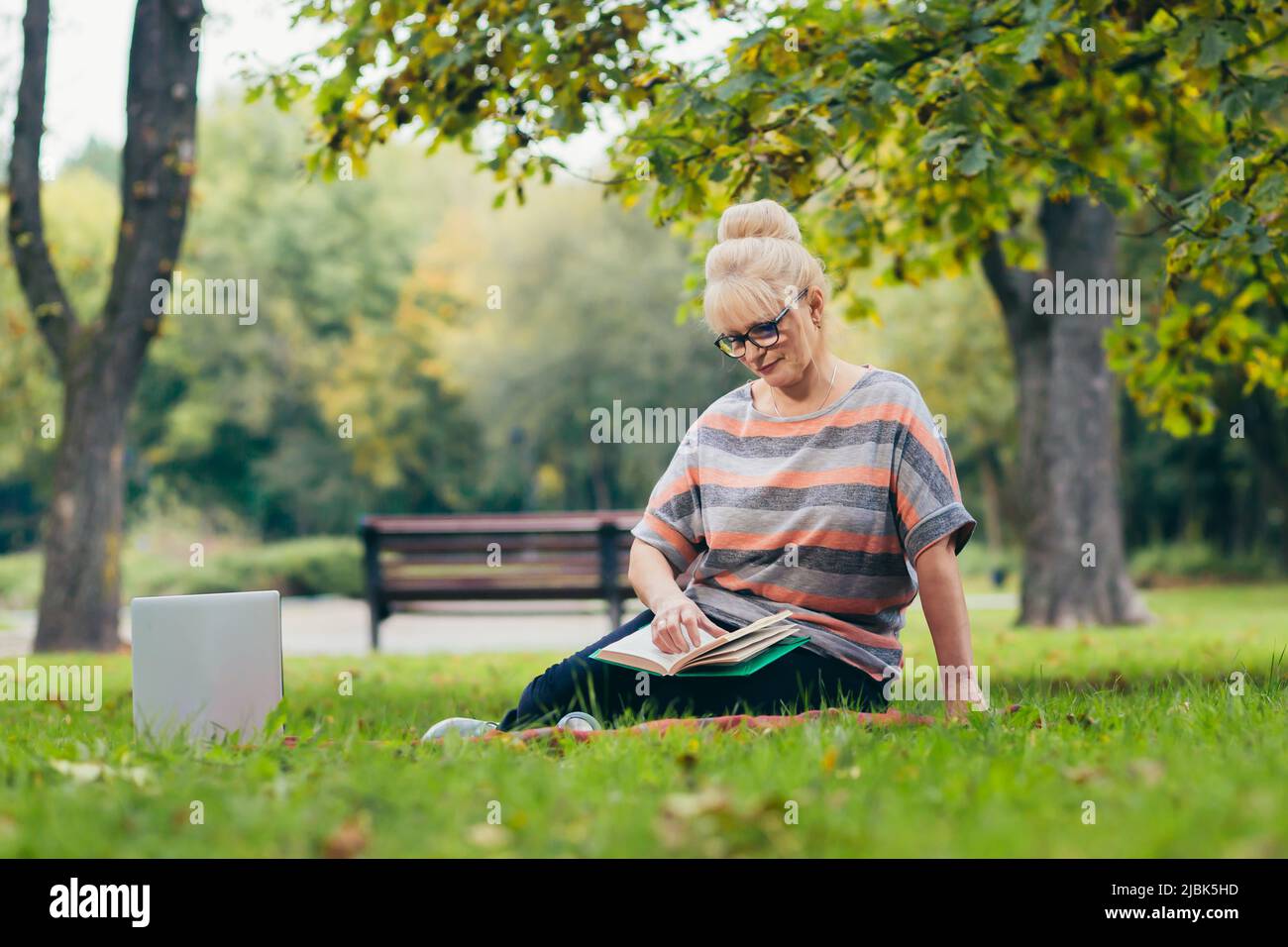 Donna bionda senior bella seduta sul prato nel parco legge un libro, studi, sorrisi, riposa Foto Stock