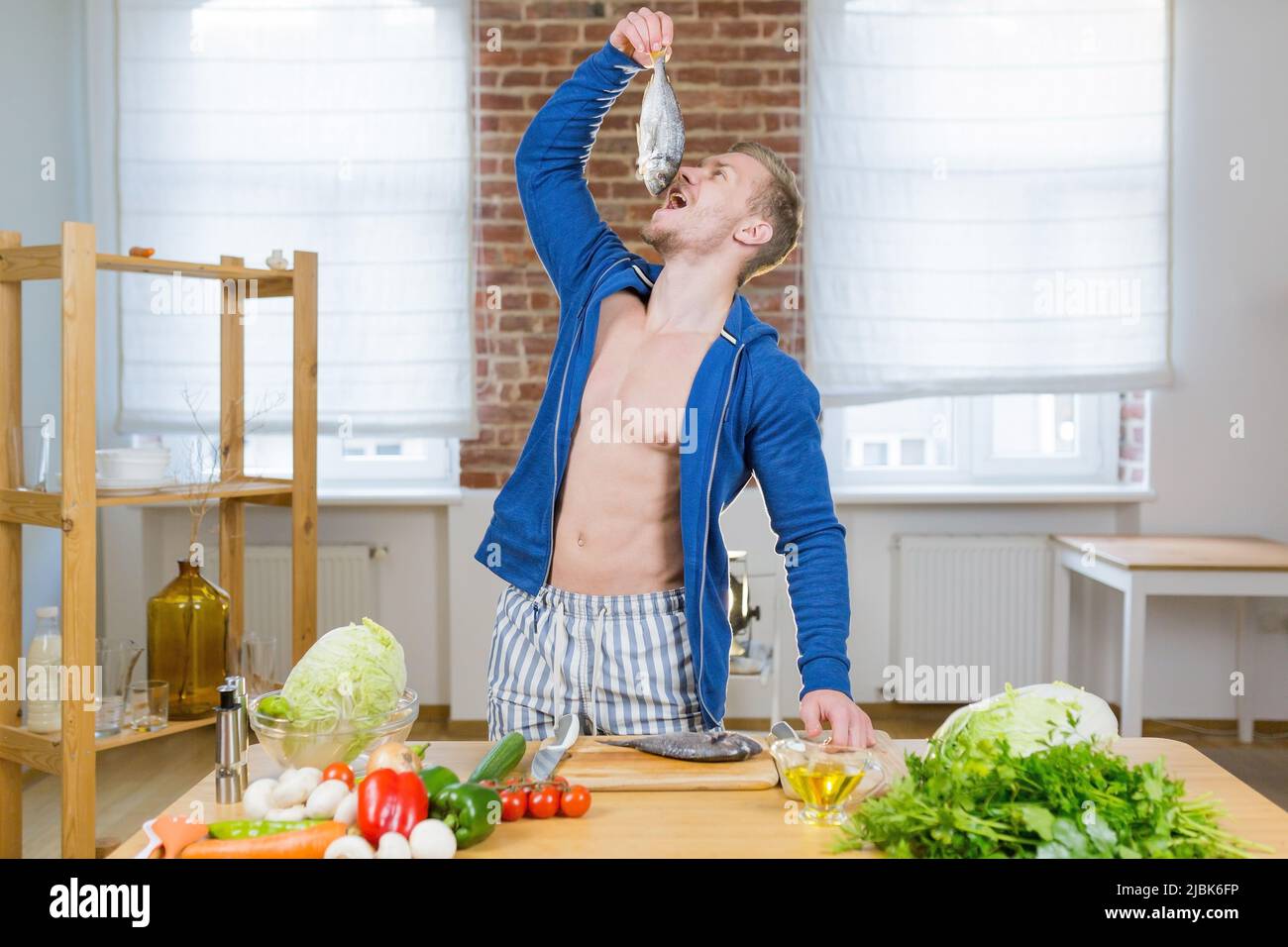 Ritratto di giovane bell'uomo atletico imparare a cucinare il pesce con verdure fresche online con il portatile in cucina a casa, guardando la macchina fotografica, sorridente Foto Stock