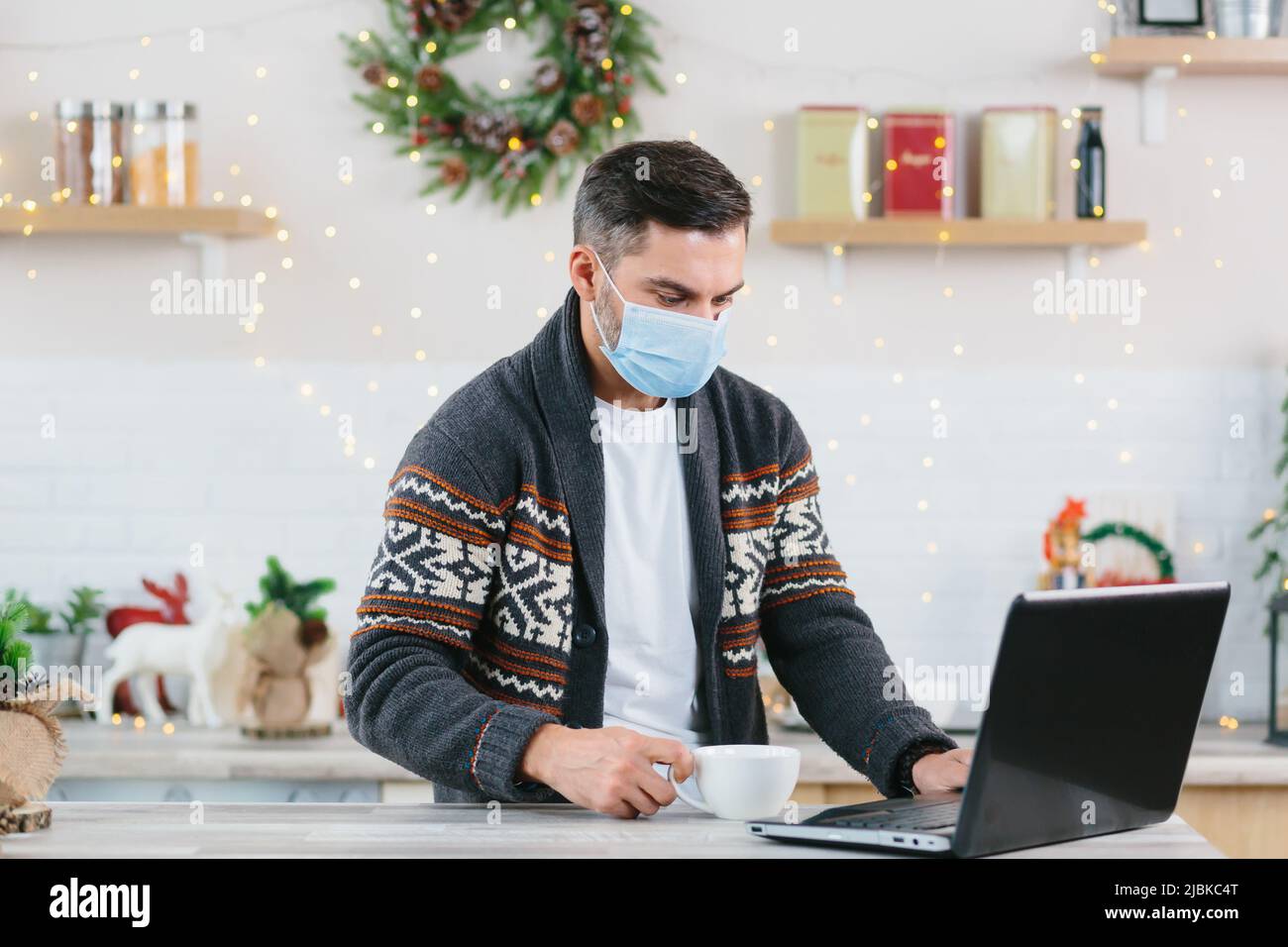 Uomo malato con un freddo in una maschera di protezione che lavora a distanza da casa utilizzando un computer portatile durante il Capodanno e le vacanze di Natale Foto Stock