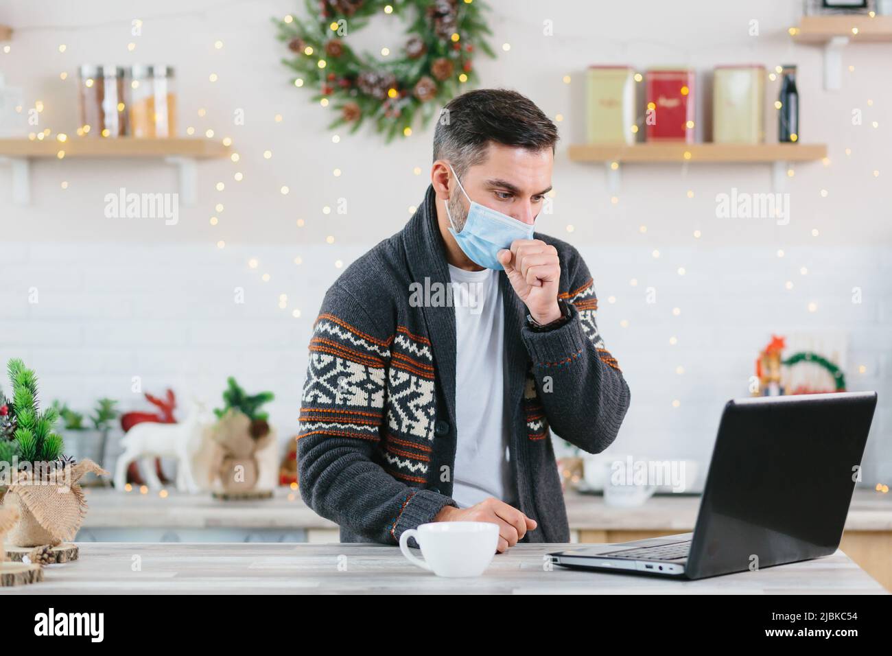 Uomo malato con un freddo in una maschera di protezione che lavora a distanza da casa utilizzando un computer portatile durante il Capodanno e le vacanze di Natale Foto Stock