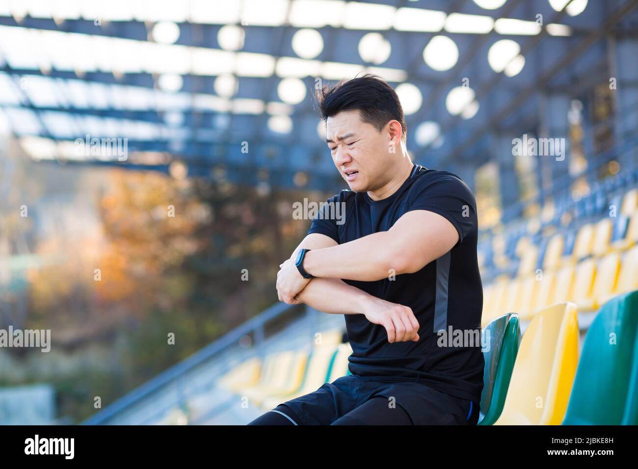 L'atleta ha un forte dolore nel braccio, l'uomo asiatico siede su una sedia nello stadio, gli inginocchiò il braccio, ferito durante l'allenamento al mattino fuori, riposandosi Foto Stock