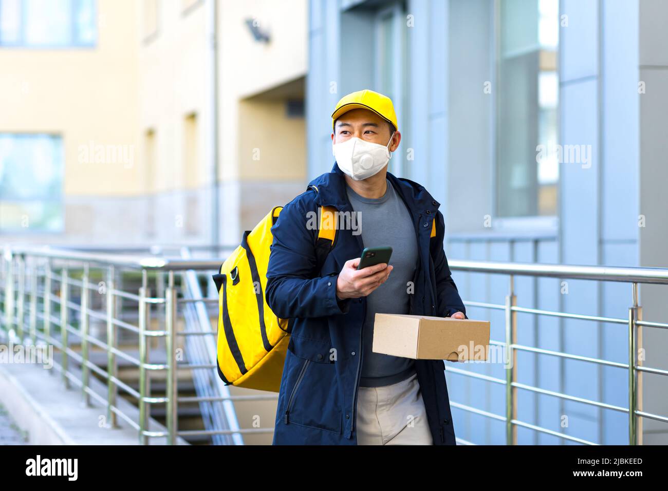 Corriere alimentare maschile asiatico in uniforme gialla utilizzando l'app mobile su smartphone mentre si cammina lungo la strada della città con isolamento con zaino termico deliverin Foto Stock