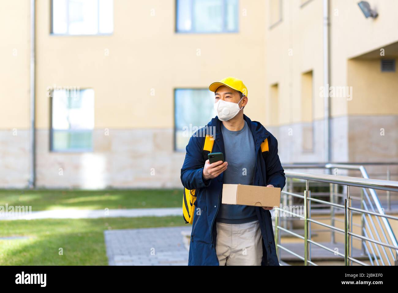 Corriere alimentare maschile asiatico in uniforme gialla utilizzando l'app mobile su smartphone mentre si cammina lungo la strada della città con isolamento con zaino termico deliverin Foto Stock