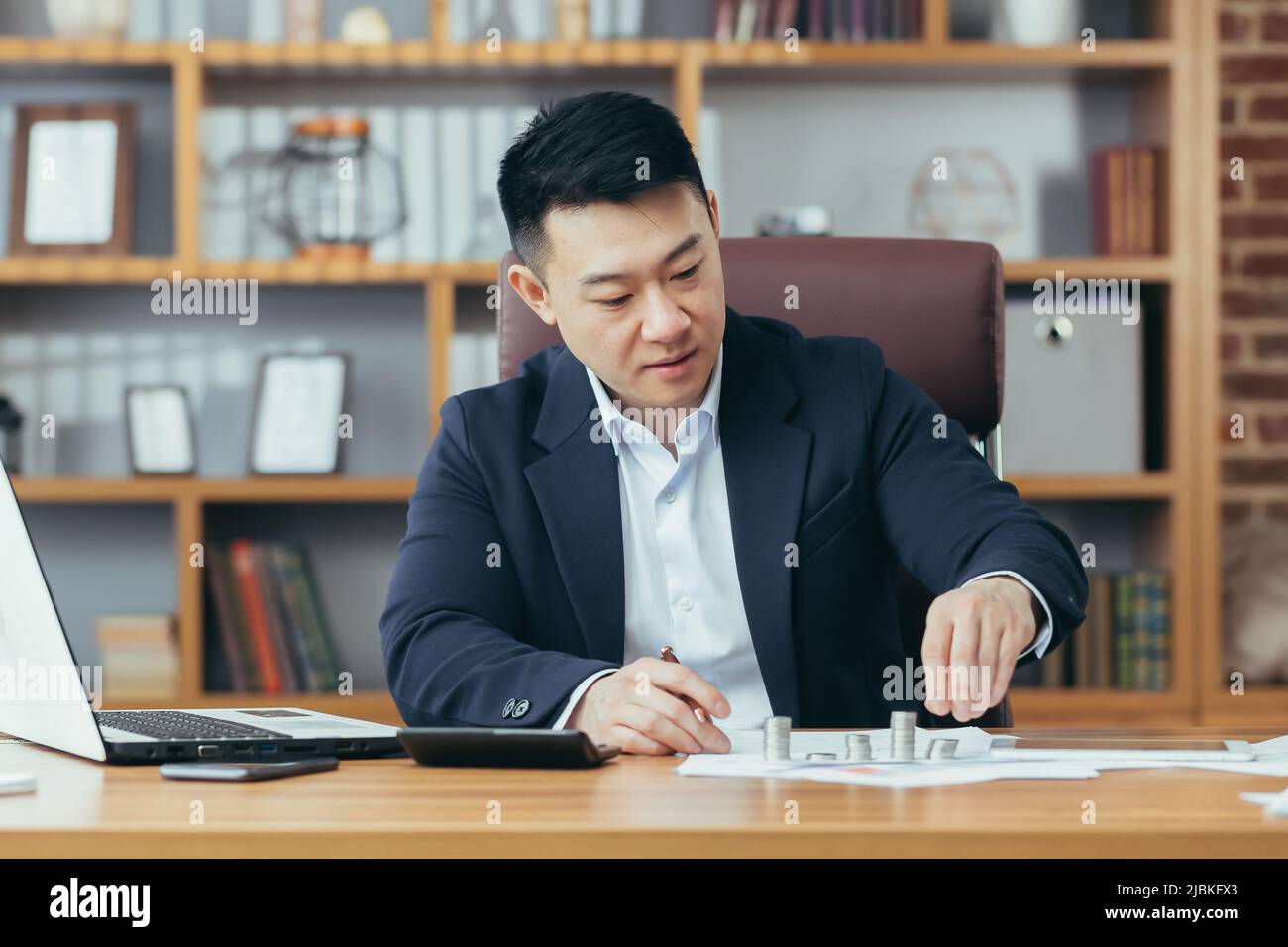 Un banchiere asiatico lavora in un ufficio classico al tavolo, contando monete in metalli preziosi Foto Stock