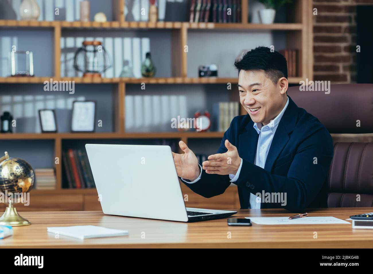 successo e felice boss asiatico guarda lo schermo del laptop e sorride, videochiamata con i colleghi, racconta piani finanziari, business meeting online Foto Stock