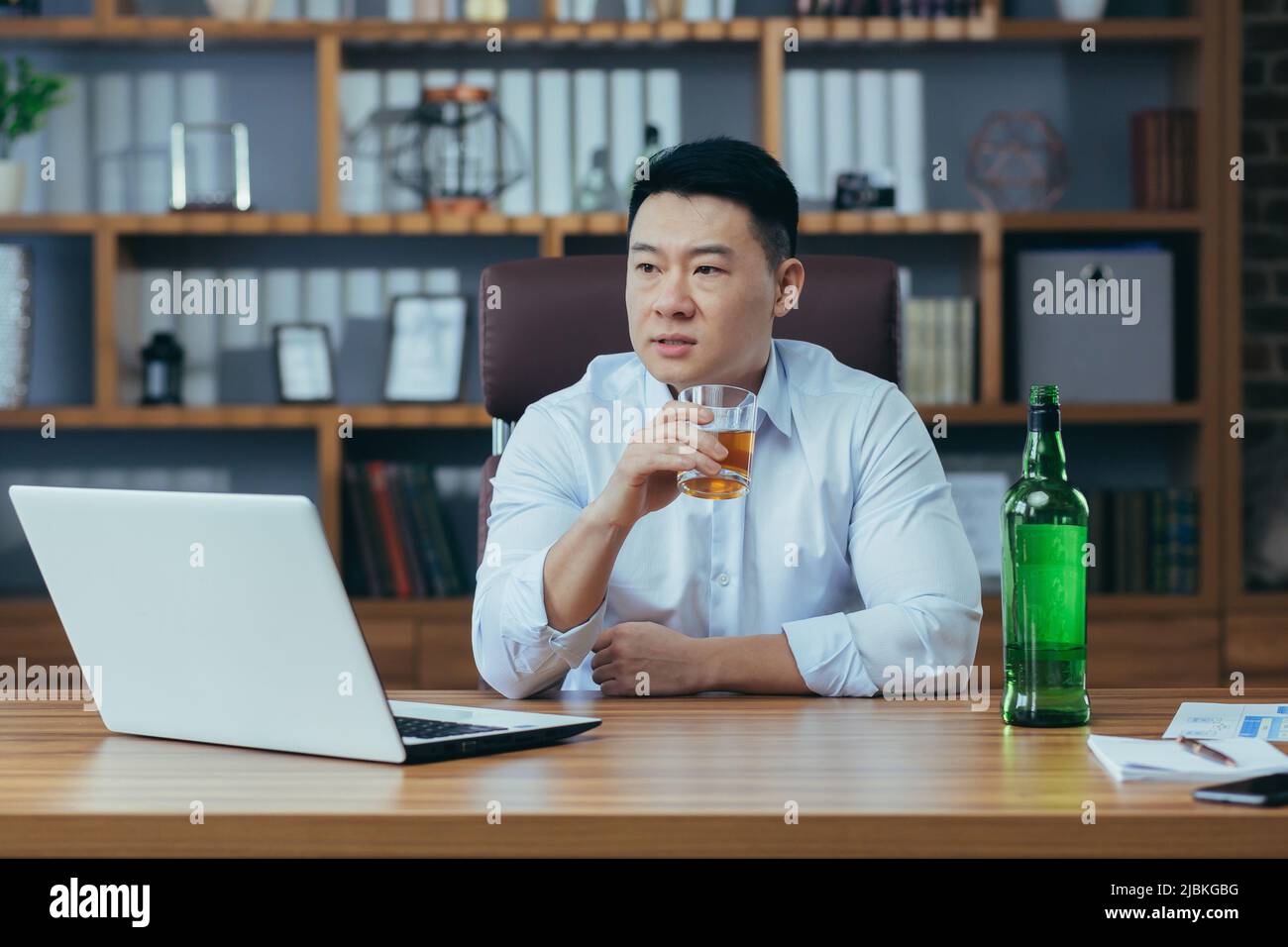 L'uomo d'affari asiatico fallisce, l'uomo disperato beve liquore duro, seduto tardi al tavolo in ufficio, depresso e senza speranza Foto Stock