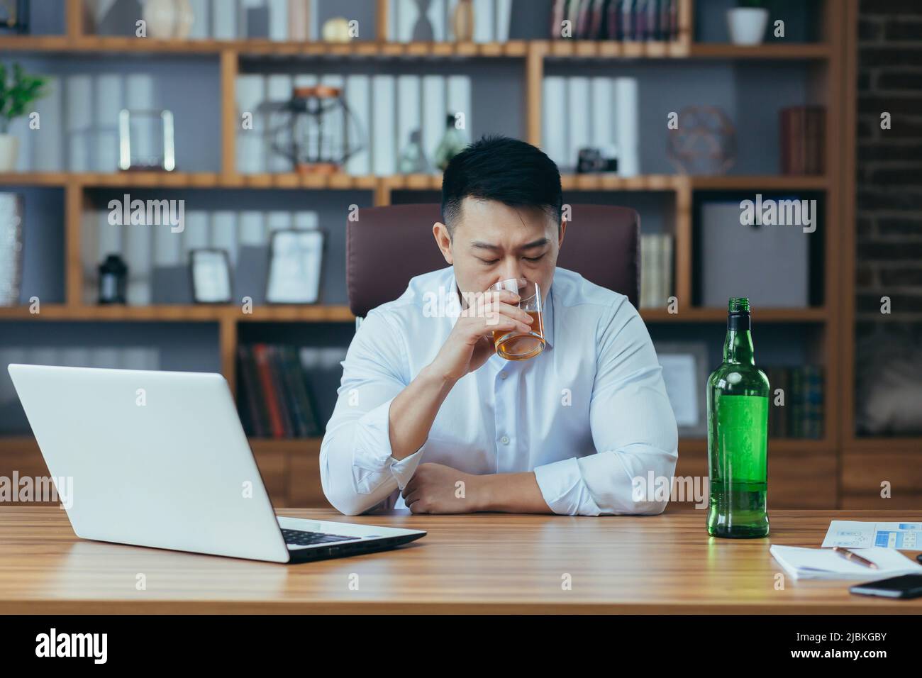 L'uomo d'affari asiatico fallisce, l'uomo disperato beve liquore duro, seduto tardi al tavolo in ufficio, depresso e senza speranza Foto Stock