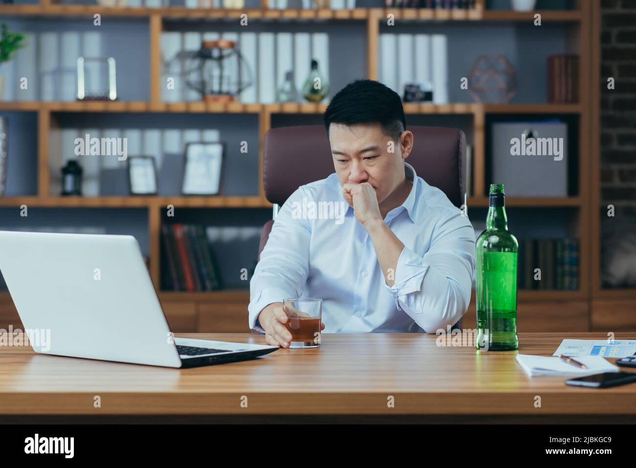 L'uomo d'affari asiatico fallisce, l'uomo disperato beve liquore duro, seduto tardi al tavolo in ufficio, depresso e senza speranza Foto Stock
