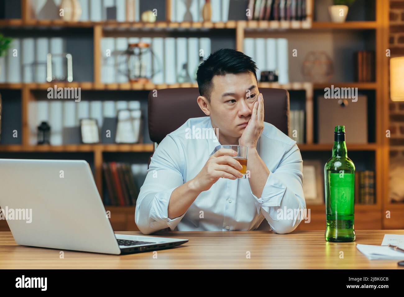 L'uomo d'affari asiatico fallisce, l'uomo disperato beve liquore duro, seduto tardi al tavolo in ufficio, depresso e senza speranza Foto Stock