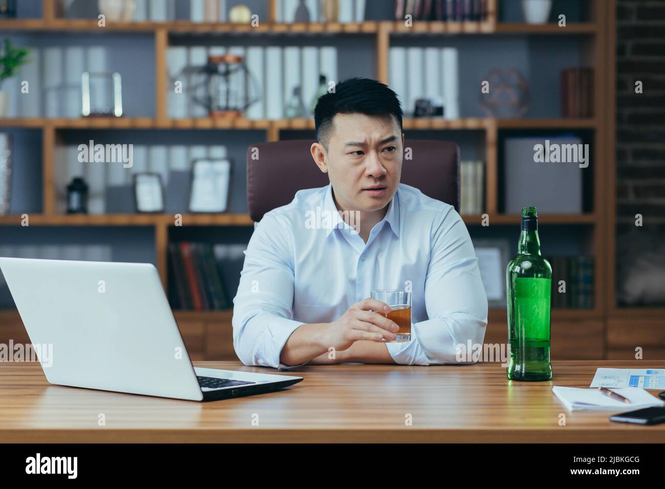 L'uomo d'affari asiatico fallisce, l'uomo disperato beve liquore duro, seduto tardi al tavolo in ufficio, depresso e senza speranza Foto Stock