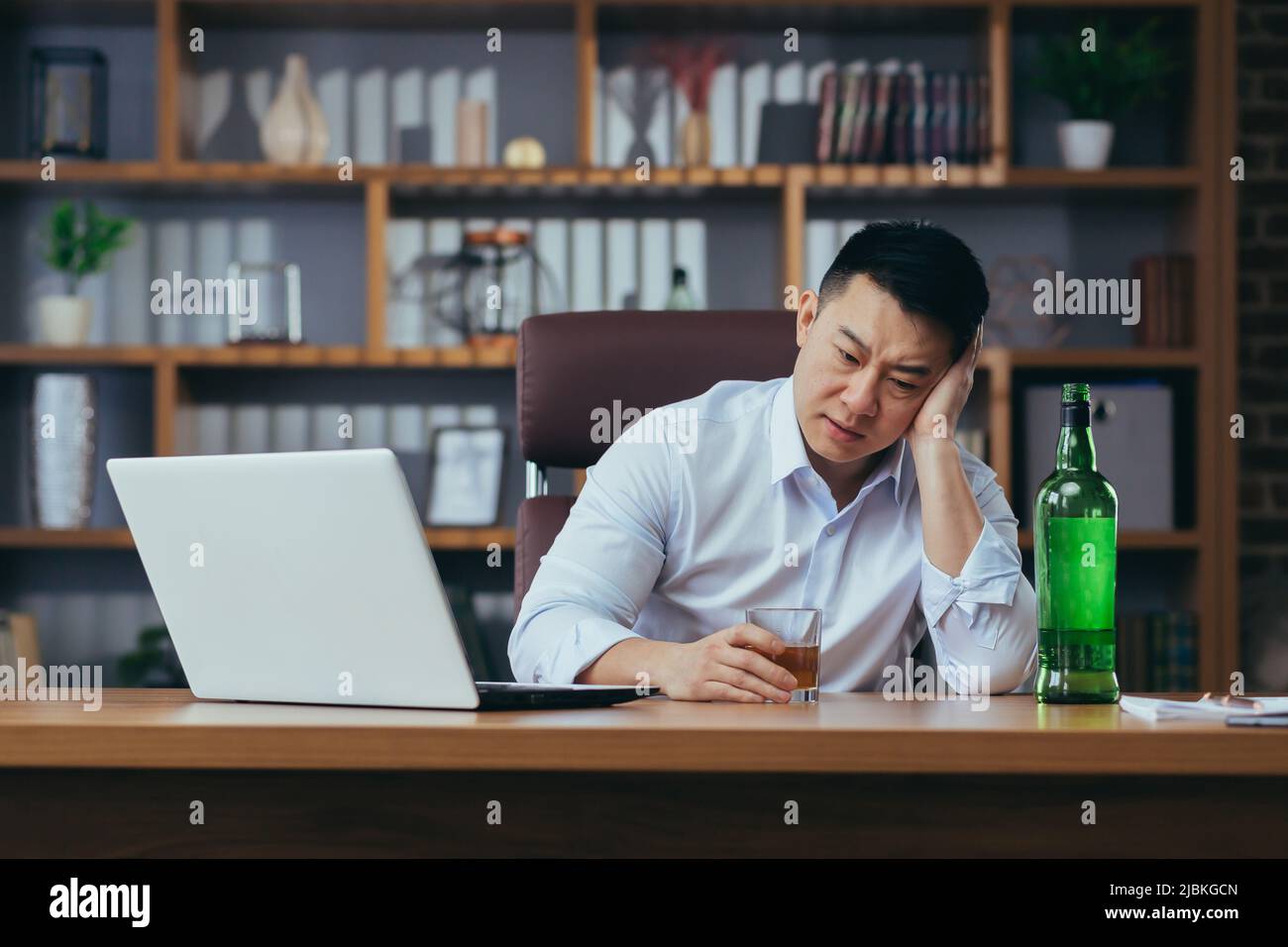 L'uomo d'affari asiatico fallisce, l'uomo disperato beve liquore duro, seduto tardi al tavolo in ufficio, depresso e senza speranza Foto Stock