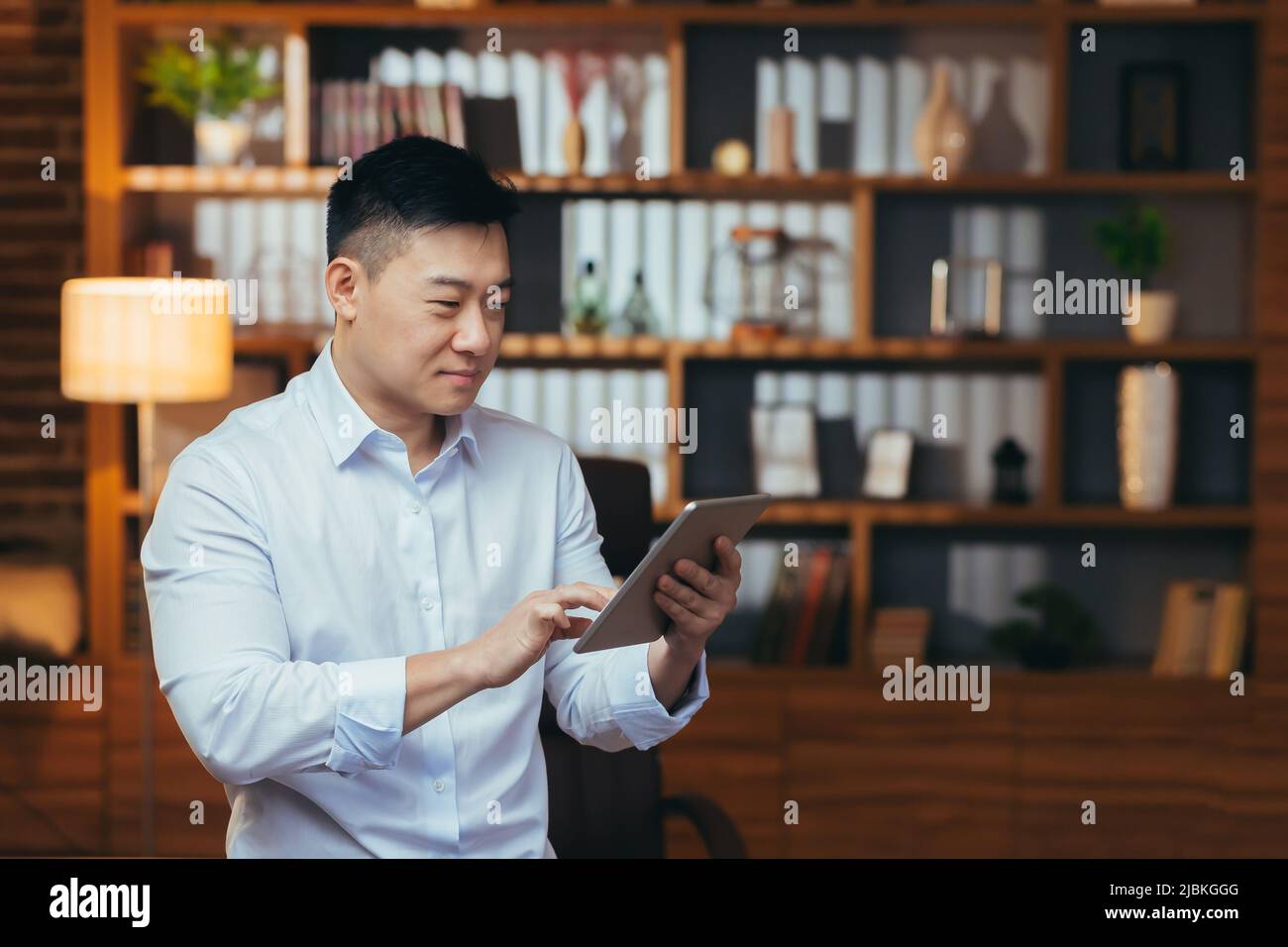 Uomo d'affari che lavora sul tablet in serata, l'uomo guarda lo schermo e sorride, lavora tardi in ufficio, felice asiatico corrisposto su un sito di incontri Foto Stock