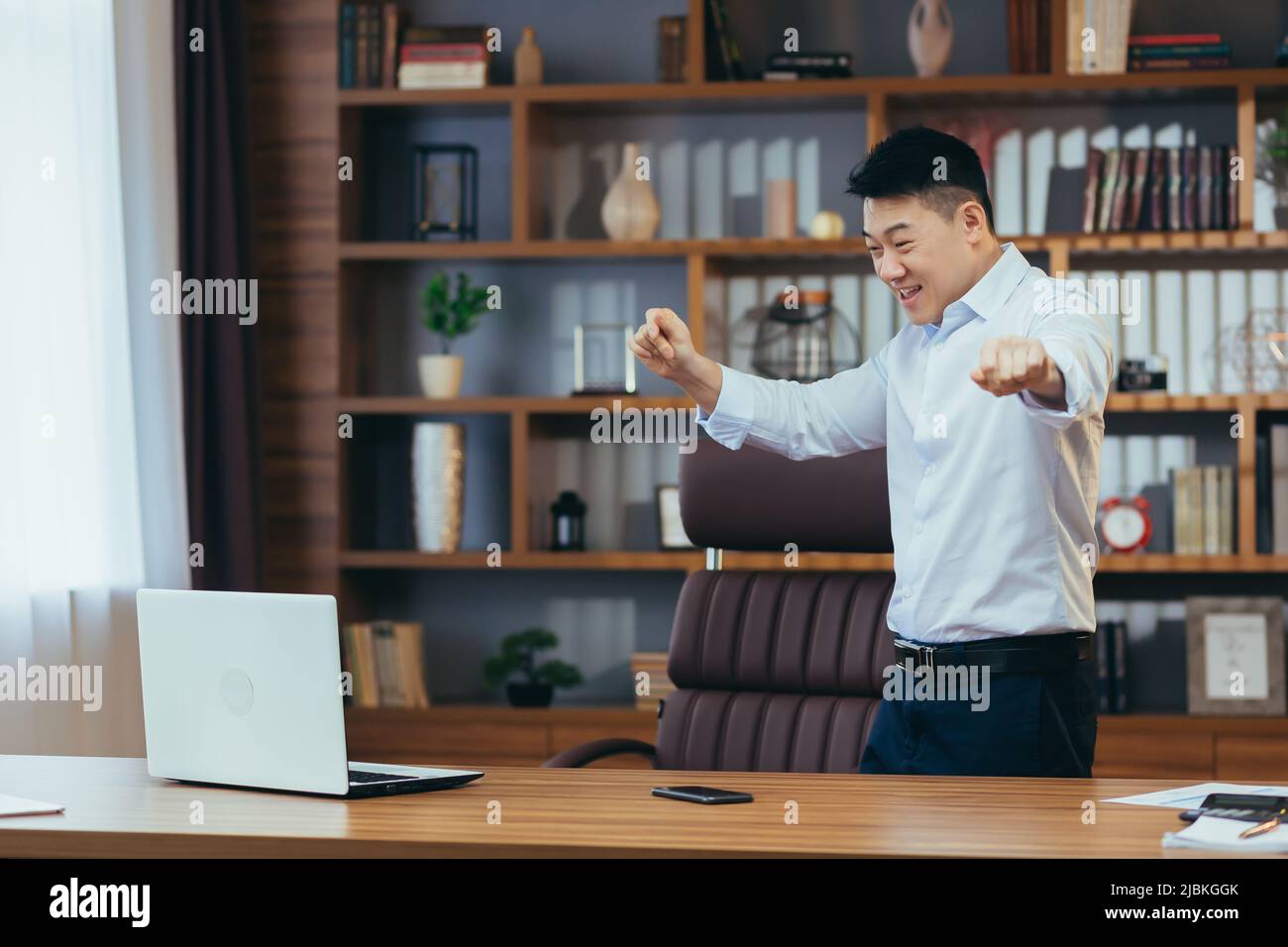 Felice uomo d'affari che balla e celebra la vittoria seduta ad un tavolo in un ufficio classico, asiatico che lavora al computer Foto Stock