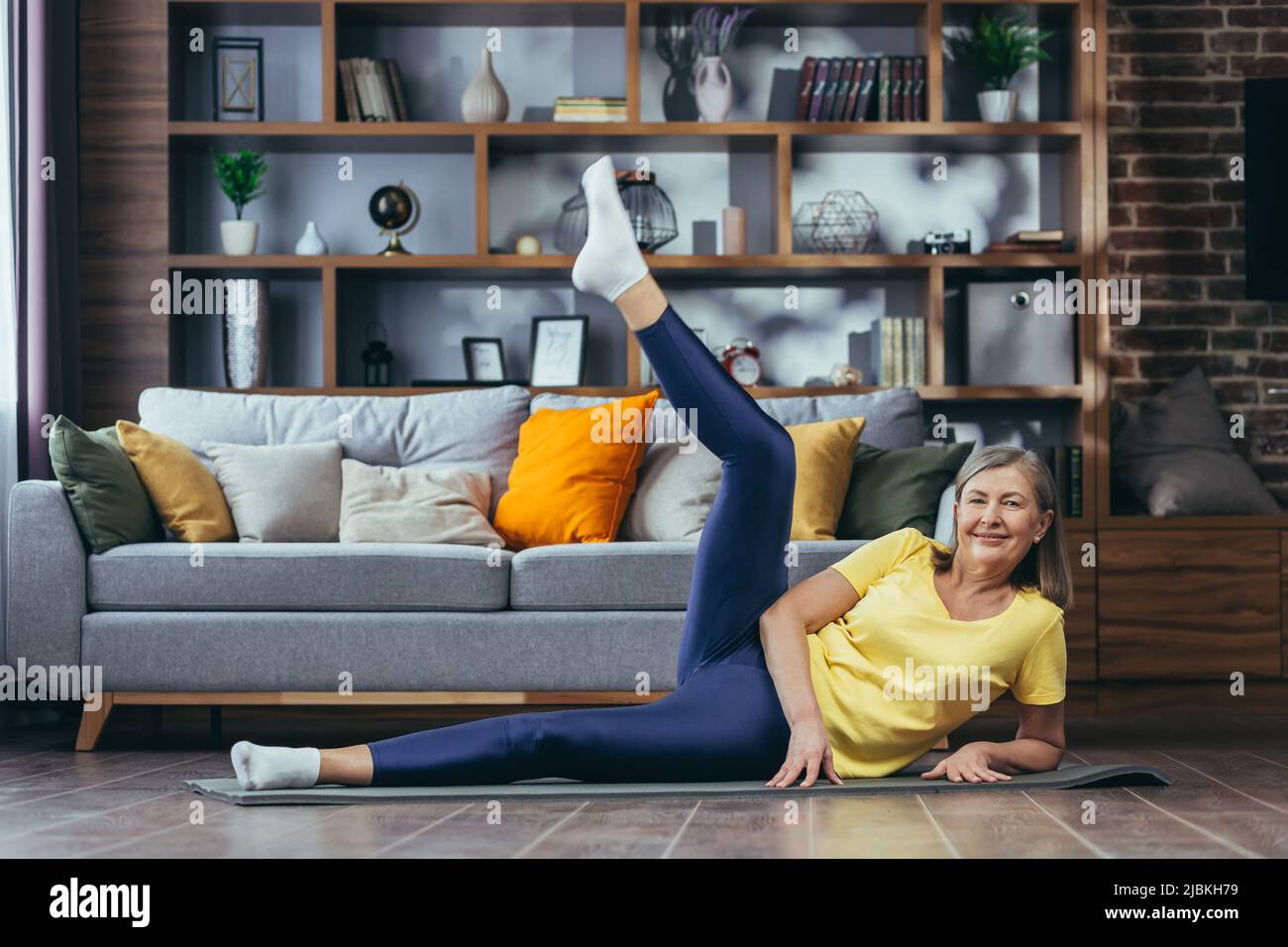 Donna anziana dai capelli grigi che fa la forma fisica a casa, facendo le esercitazioni del mattino su un tappeto sportivo seduto sul pavimento a casa Foto Stock