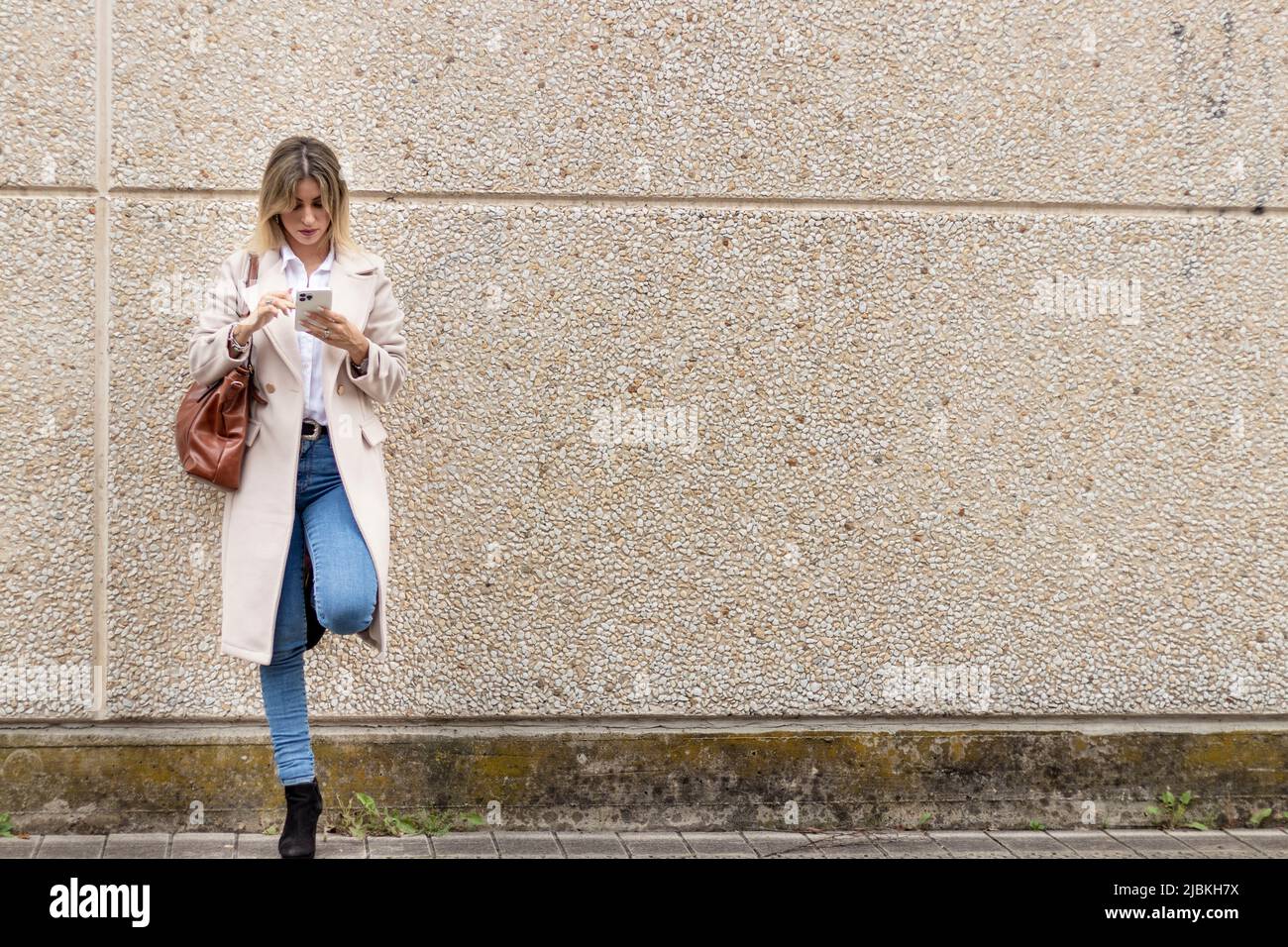 donna in cappotto beige, camicia bianca e jeans appoggiati alla parete guardando il mobile Foto Stock