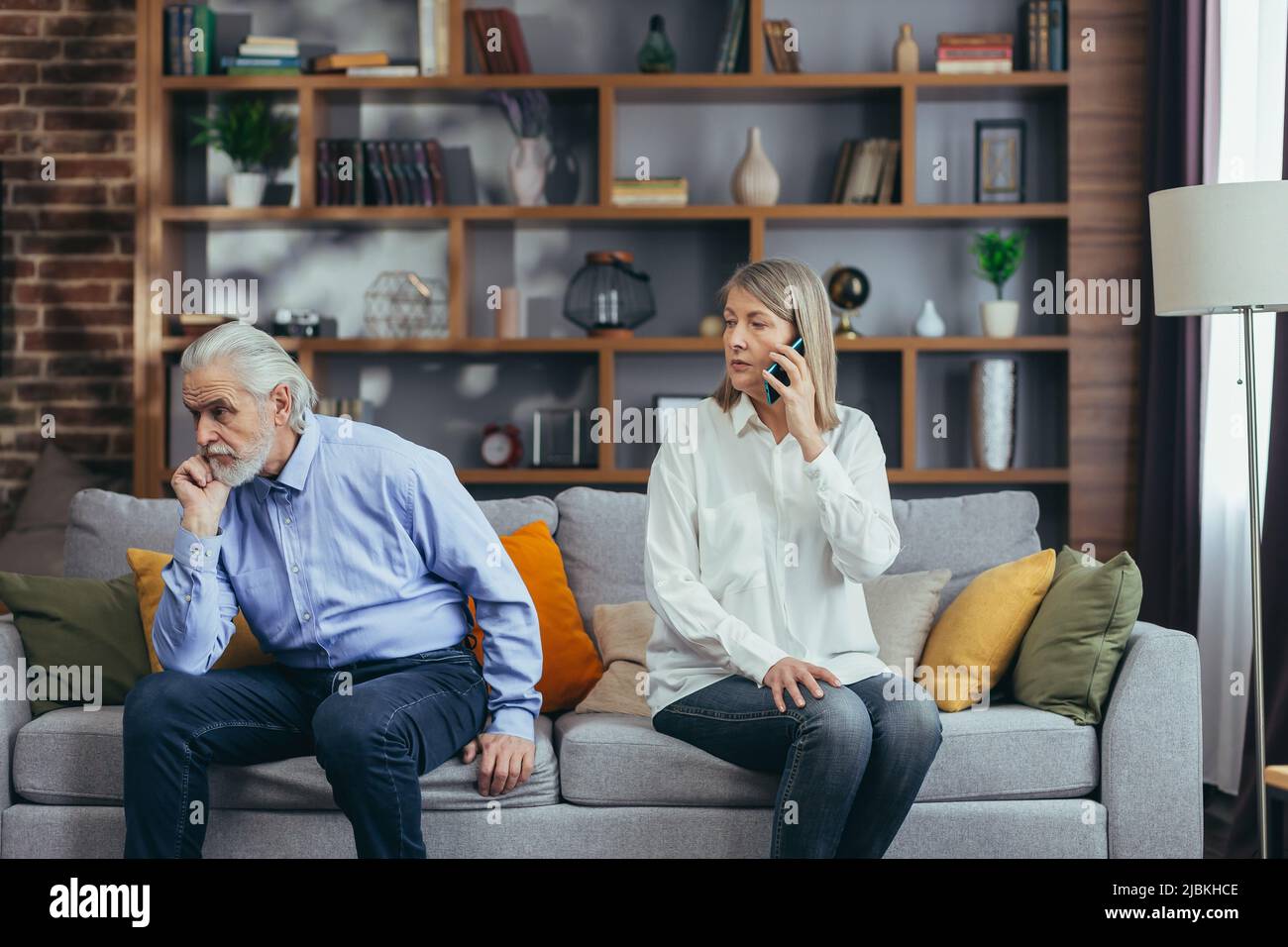 coppia sposata senior, litigante, uomo e donna dai capelli grigi seduti sul divano, moglie infastidita a parlare al telefono Foto Stock