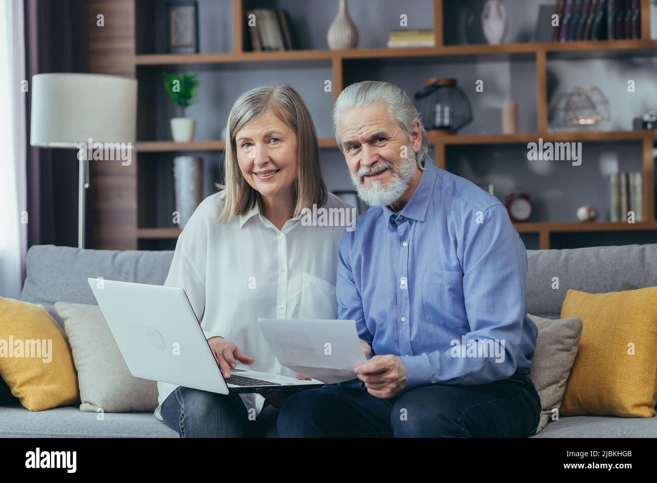 Senior grigio-capelli sposati uomo e donna coppia seduta insieme sul divano a casa, guardando la macchina fotografica e sorridente, carta lavoro con il portatile Foto Stock