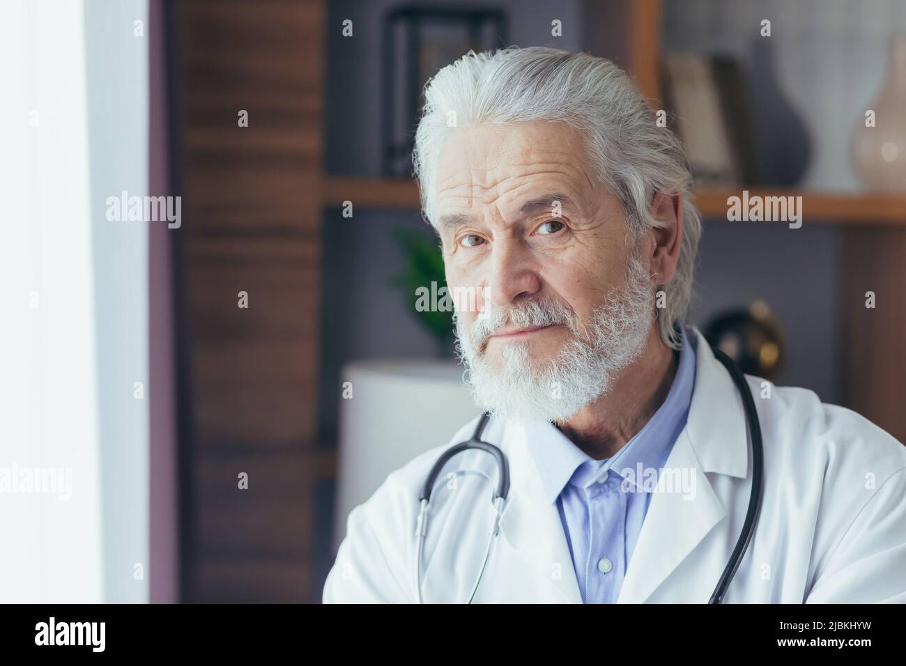 ritratto di primo piano di un medico esperto, un uomo dai capelli grigi che guarda nella macchina fotografica, lavora in un ufficio privato, vestito in un medico Foto Stock