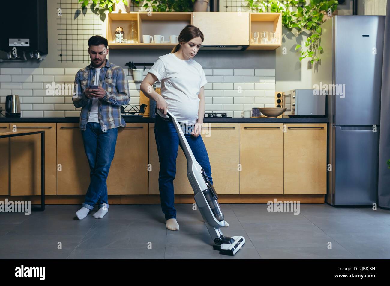 Famiglia a casa, donna incinta pulisce la stanza con un aspirapolvere, marito usa il telefono in cucina Foto Stock