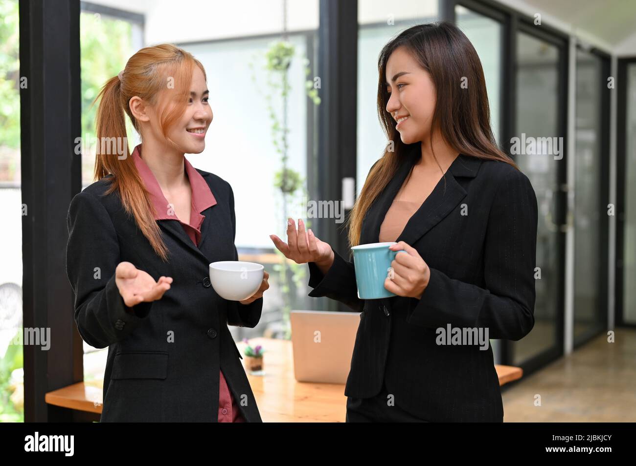 Due belle e di successo donne d'affari asiatiche in tuta formale in piedi in ufficio, avendo un caffè e godere di chiacchierare insieme durante il caffè Foto Stock