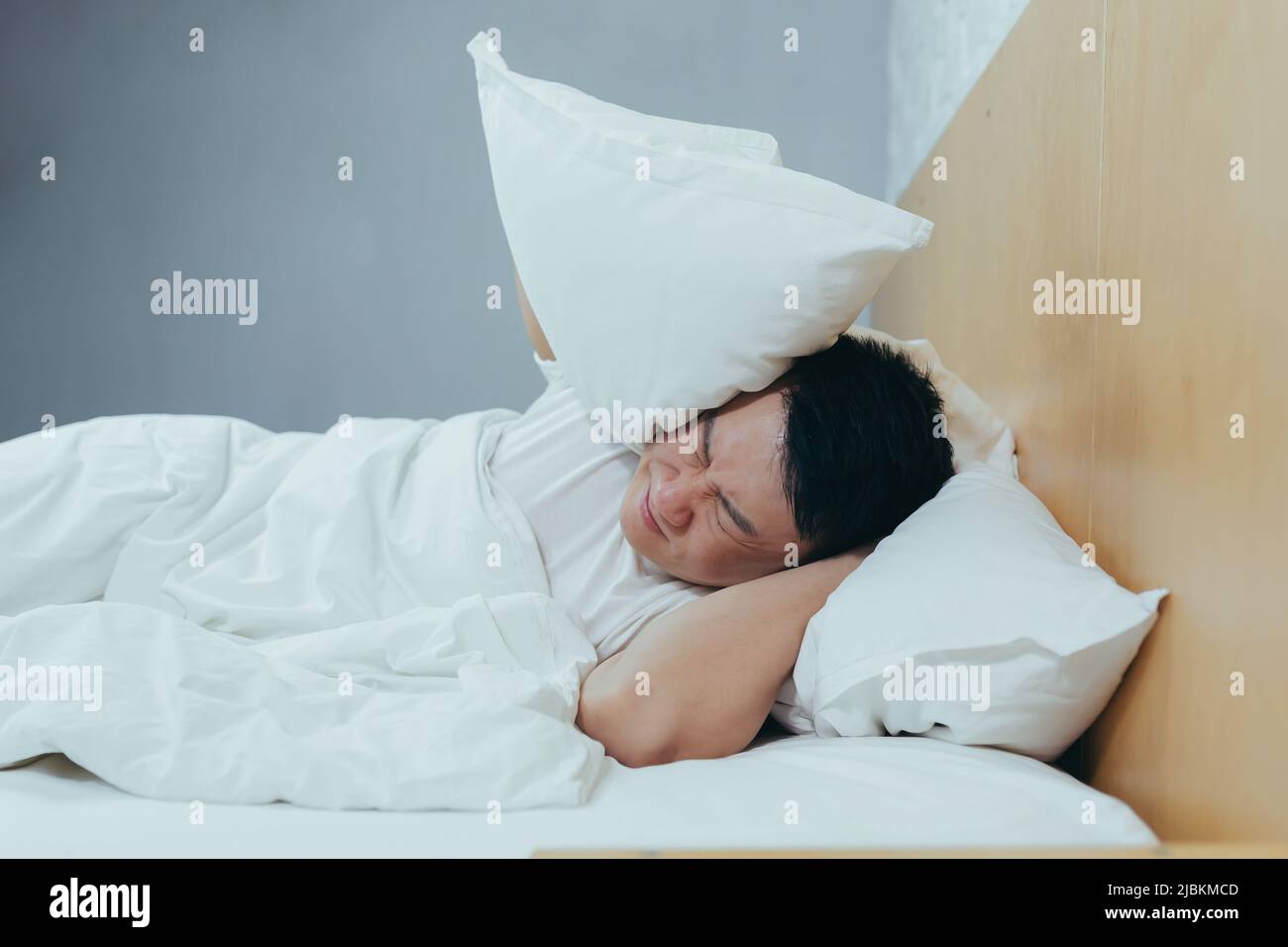 Un uomo a casa cerca di addormentarsi, vicini rumorosi interferiscono con il sonno, un asiatico chiude le orecchie con cuscini, stanchi dopo il lavoro Foto Stock