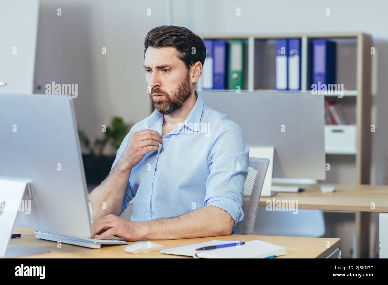 Caldo in ufficio, un uomo d'affari in una camicia che lavora in un luminoso ufficio moderno, agitando le braccia cercando di rinfrescarsi Foto Stock