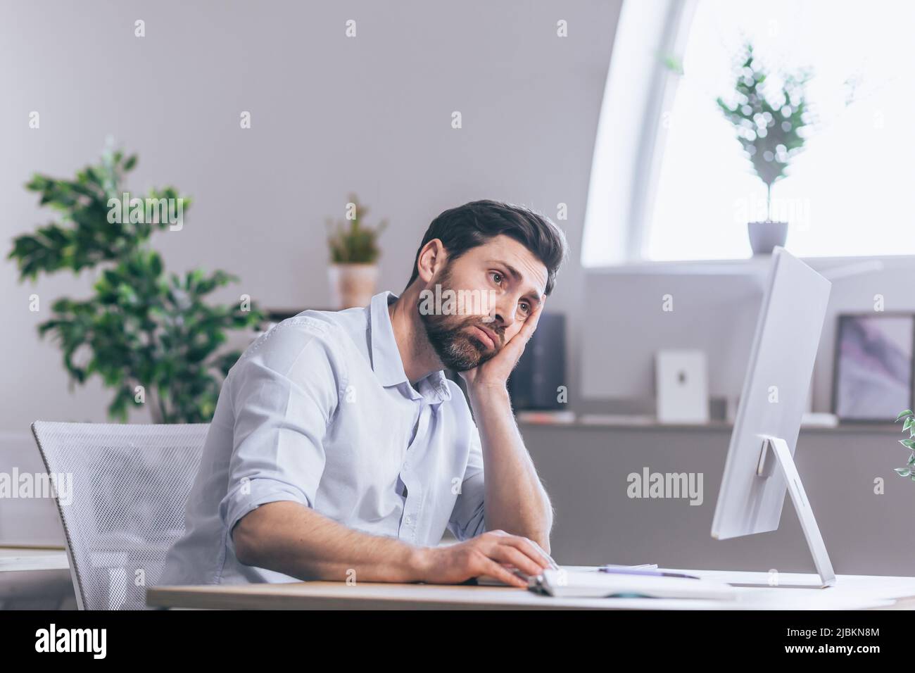 Lavoratore da ufficio stanco e attento. Seduto ad una scrivania in ufficio, al computer, tenendo la testa nelle mani, stanco, in una camicia bianca Foto Stock