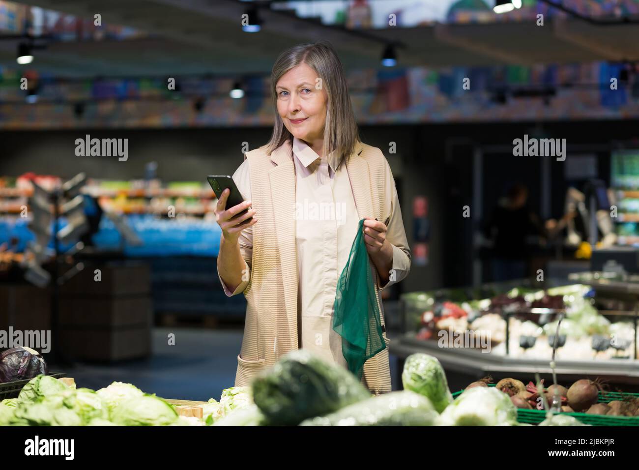 Donna anziana in un supermercato sceglie merci, guarda la lista elettronica al telefono Foto Stock