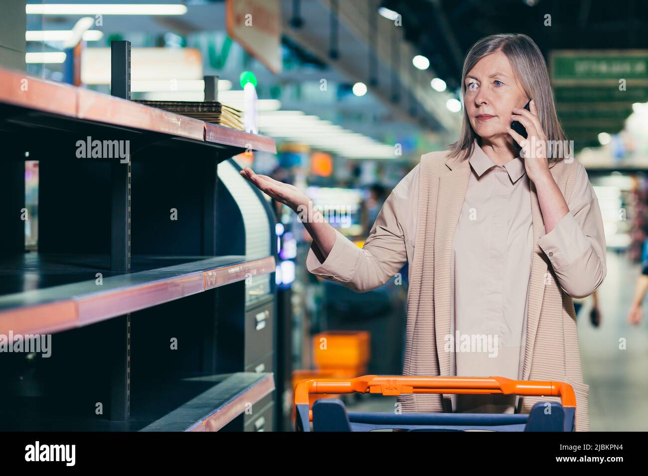 Quarantena, guerra, deficit. Donna anziana nel negozio, supermercato, parlando al telefono. Si stende le braccia, punta agli scaffali vuoti. Non ci sono Foto Stock