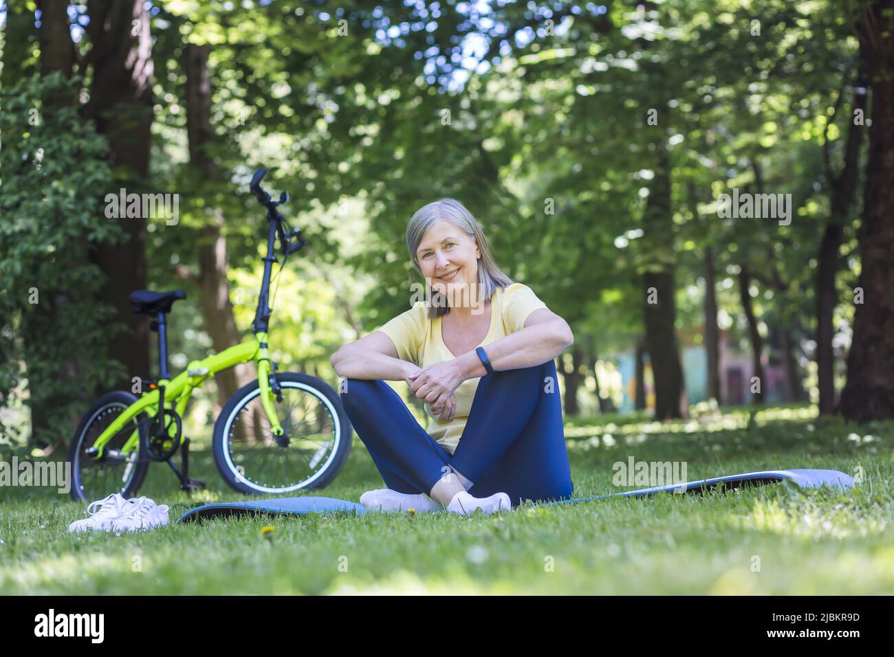 Stile di vita attivo e sano. Ritratto di una bella donna anziana. Seduto nel parco sull'erba su un tappeto sportivo con una bicicletta, guardando la ca Foto Stock