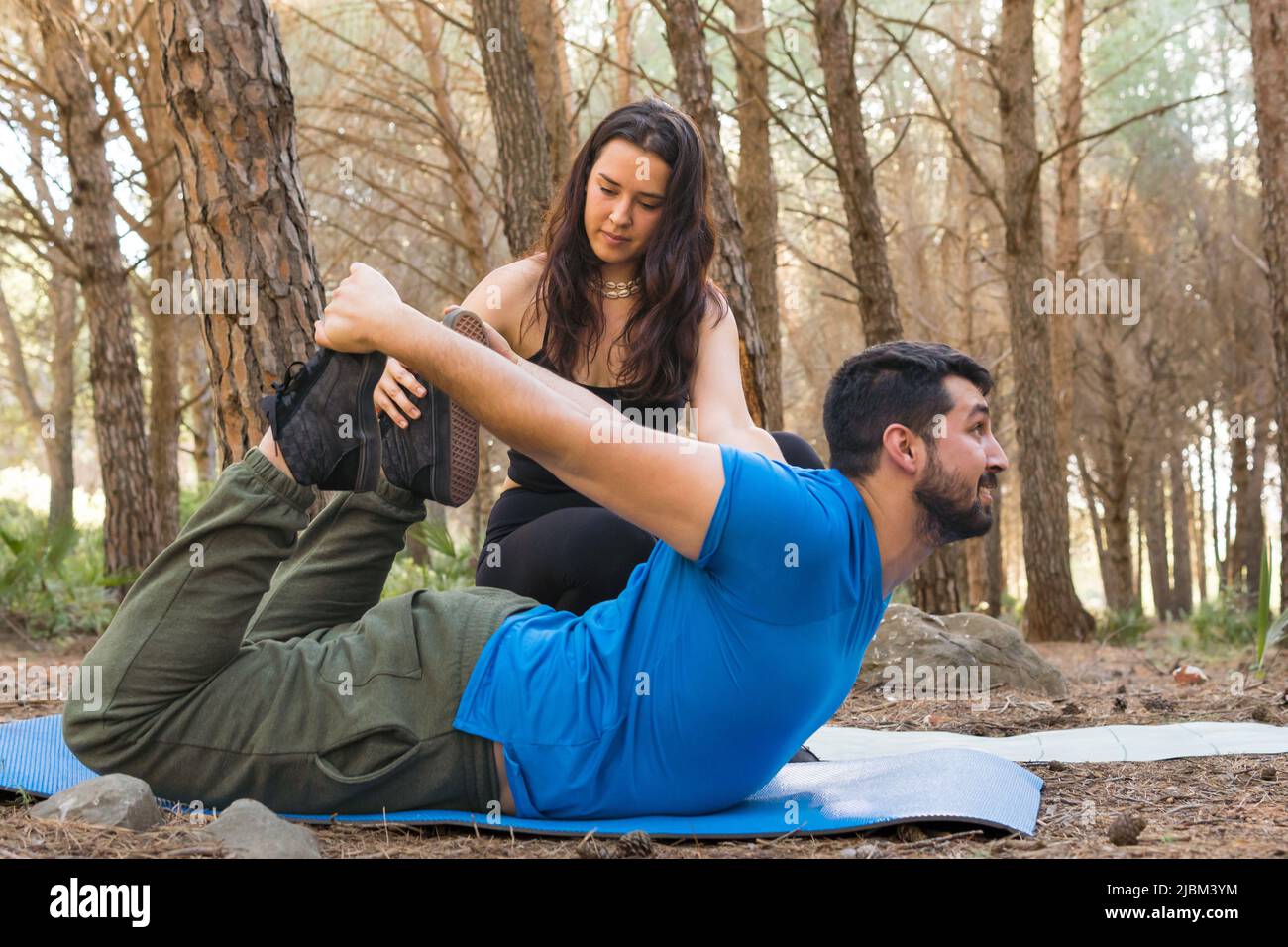 La ragazza graziosa insegna al suo amico che si trova faccia in giù sul suo tappeto per praticare yoga in una foresta al tramonto Foto Stock