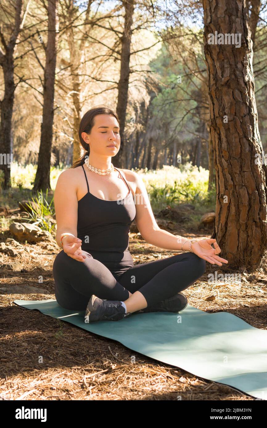 bella ragazza giovane adulta che fa meditazione nella postura del loto al tramonto in una foresta circondata da alberi. piano aperto, colpo verticale. Foto Stock